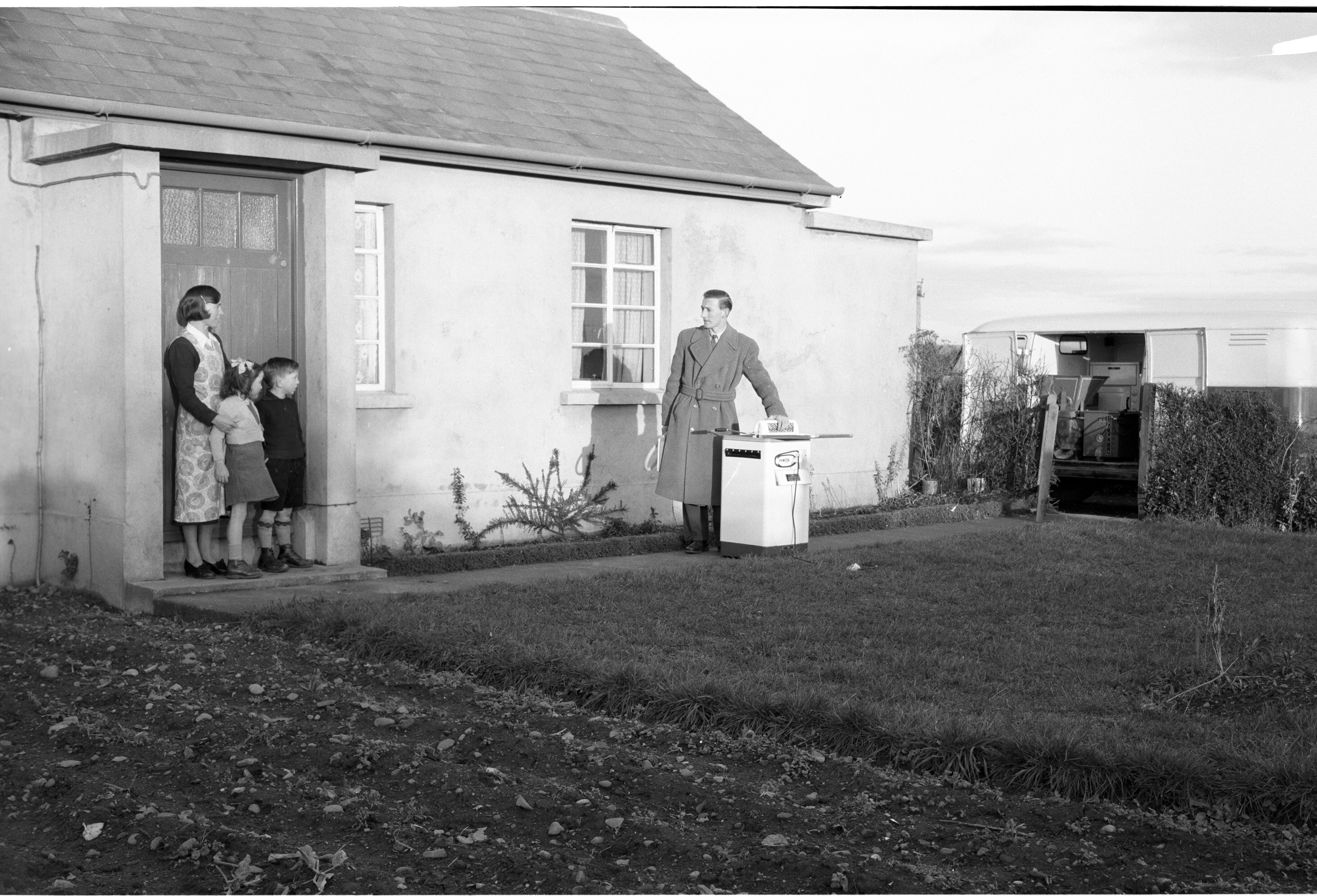 Man standing with stove outside of a cottage with woman and children looking at him from the doorway
