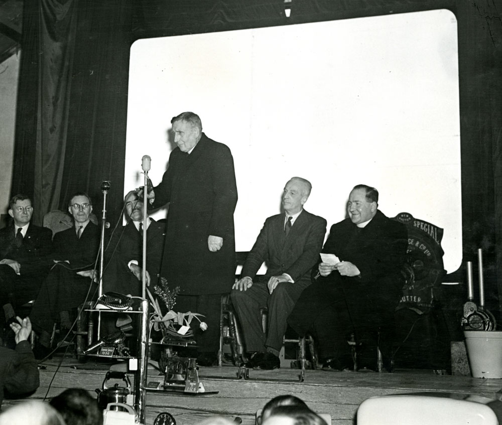 Black-and-white photograph showing a speaker standing at a microphone on a stage, with several people seated behind during a public meeting or community event.