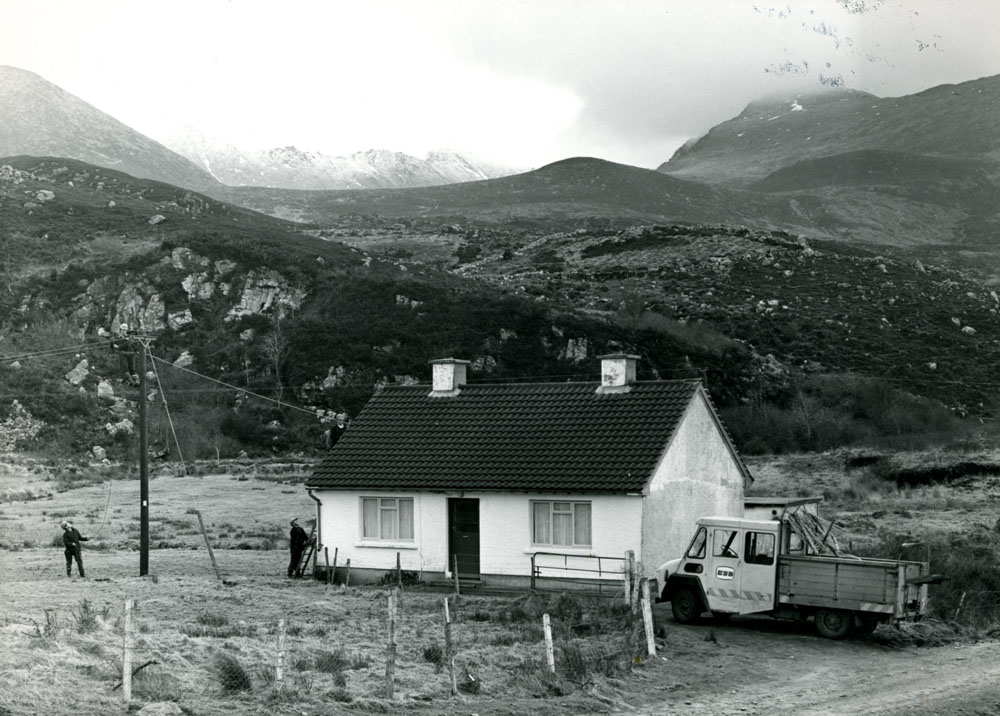 Black-and-white photograph showing an ESB crew working on an electricity service connection beside a rural house, with a utility vehicle parked nearby and mountains in the background.