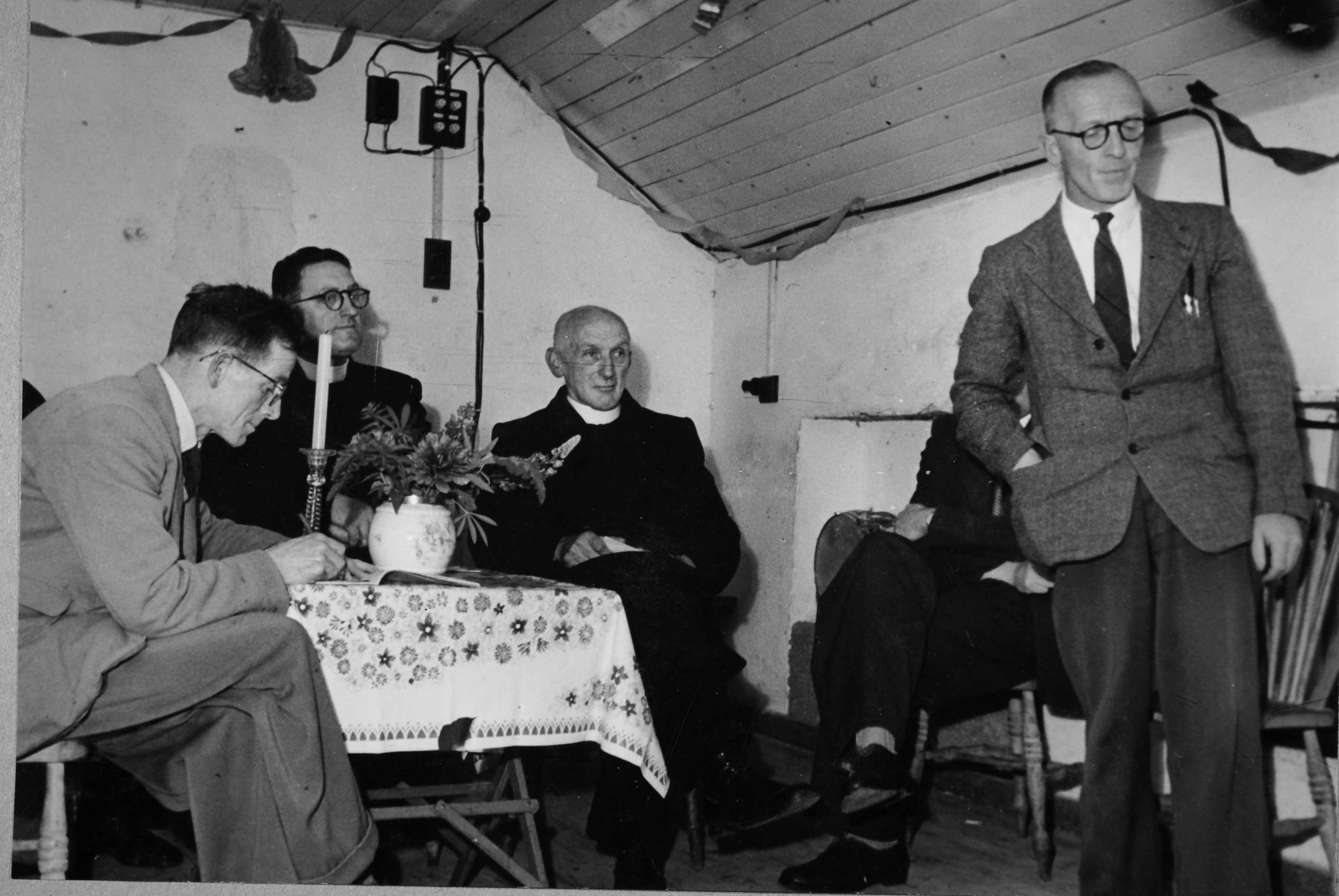 Black-and-white photograph showing a community meeting in a small hall, with a speaker standing at the front while several attendees are seated nearby.