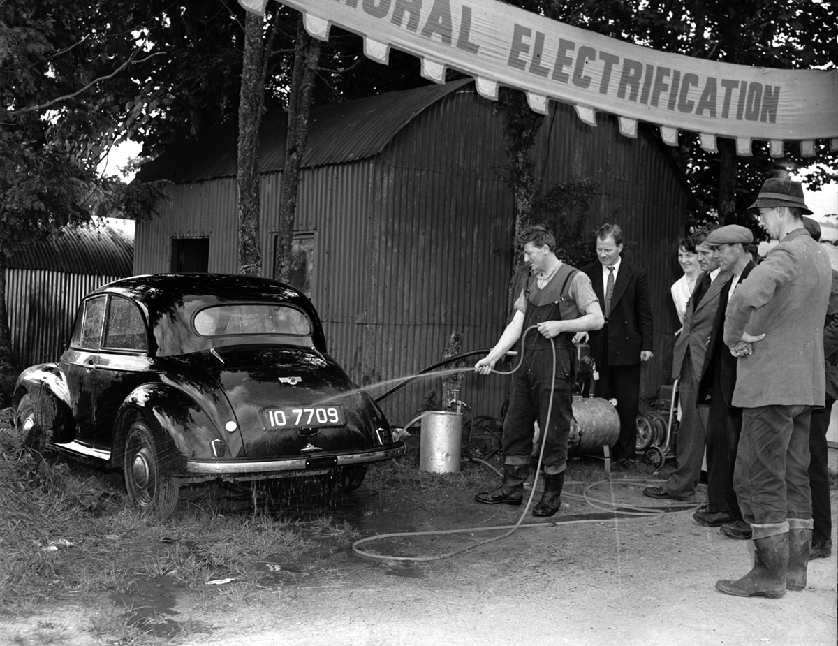 Black-and-white photograph showing a demonstration of an electrically powered pressure washer cleaning a car, with a group of onlookers gathered beside a farm building.