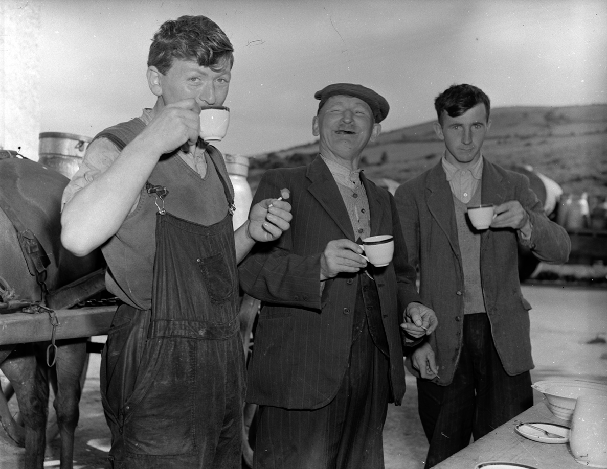 Black-and-white photograph showing several people gathered around a table drinking tea outdoors, with a horse and cart visible beside the group.