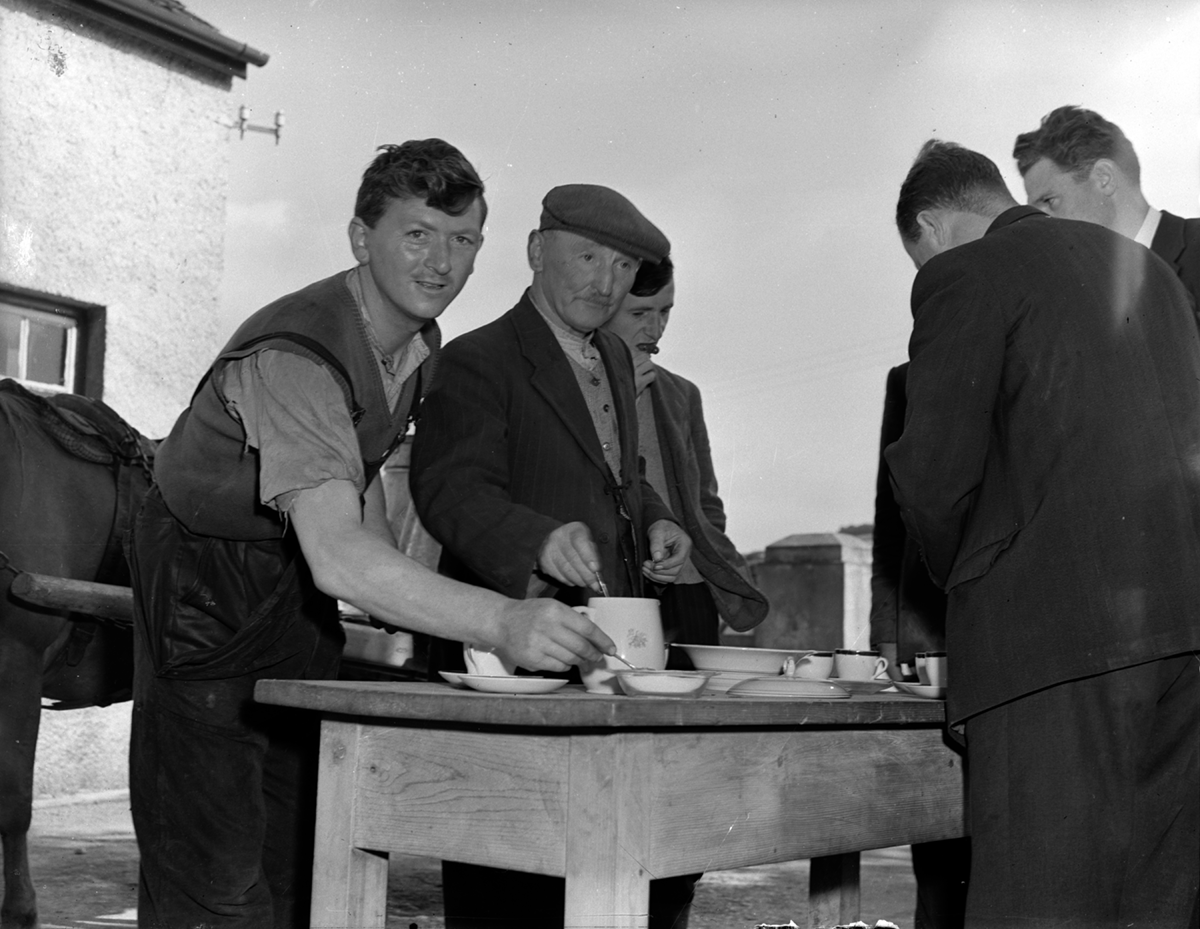Black-and-white photograph showing several people gathered around a table serving tea outdoors, with a horse and cart visible beside the group.