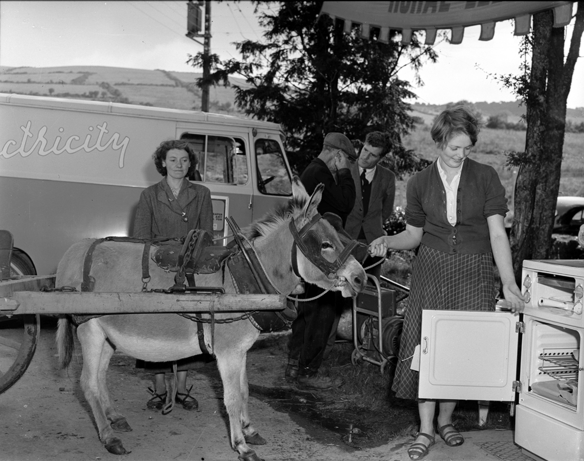 Black-and-white photograph showing a demonstration of an electric cooker beside a donkey and cart at an outdoor rural electrification event.