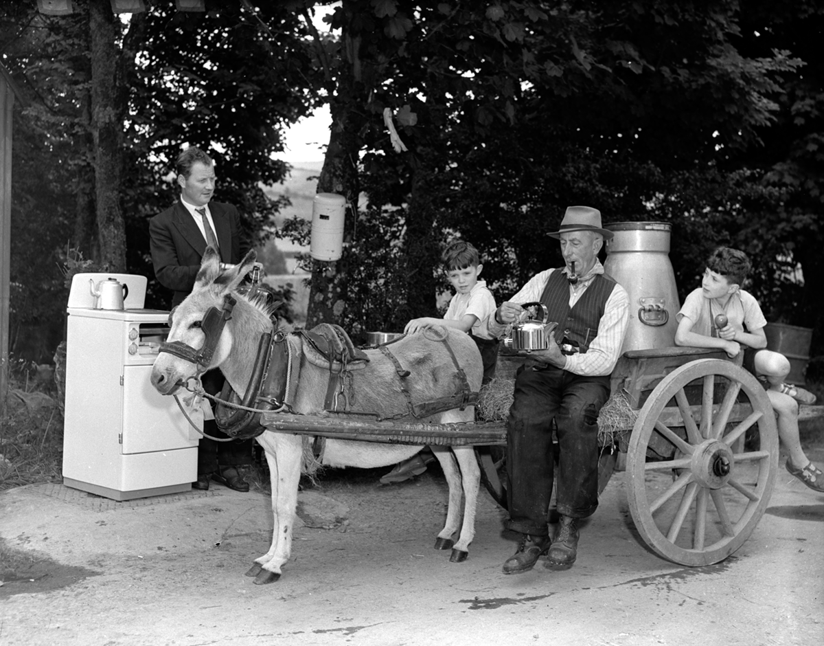Black-and-white photograph showing a donkey and cart beside a display of household electrical appliances, including an electric cooker and water heater, during a rural electrification promotion.