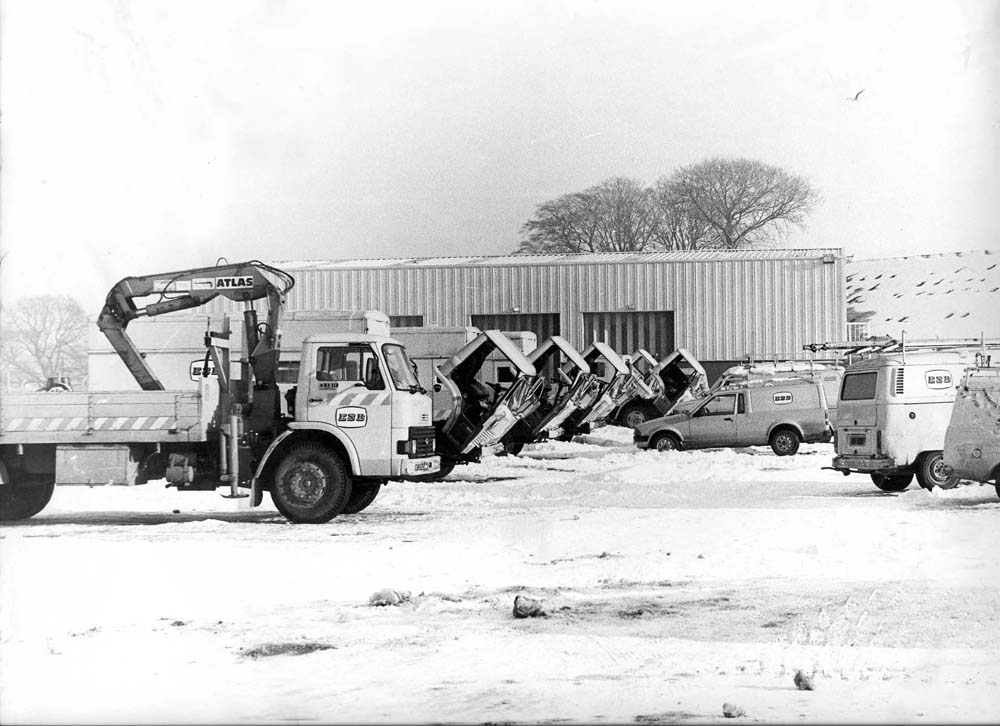 Black-and-white photograph showing ESB utility vehicles parked outside a depot in snowy conditions, including a lorry fitted with a crane and a row of vehicles with raised tipping bodies.