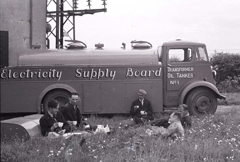 Black-and-white photograph showing an oil tanker parked beside an electricity substation while a group of workers sit in the grass having a break.