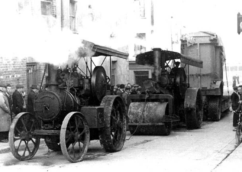 Black-and-white photograph showing steam-powered road construction machinery and a tipper lorry parked on a city street, with a crowd gathered nearby.