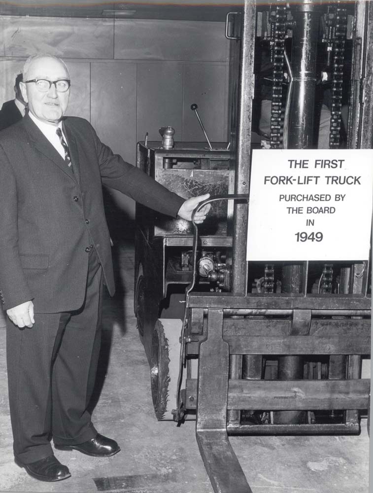 Black-and-white photograph showing a person standing beside an early forklift truck displayed as part of a company vehicle collection or exhibition.