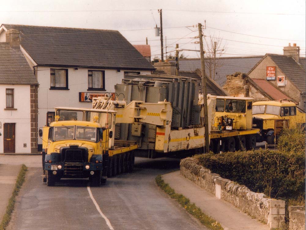 Colour photograph showing a heavy transport convoy moving a large electrical transformer through a village street, accompanied by support vehicles.