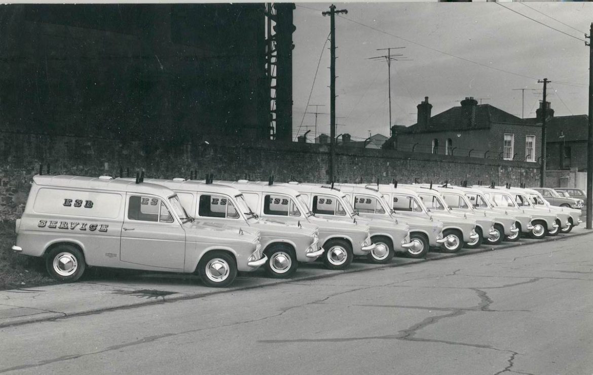 Black-and-white photograph showing a row of ESB service vans parked along a street beside an industrial wall.