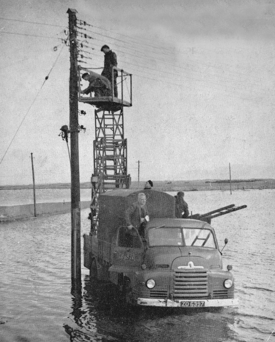 Black-and-white photograph showing an ESB utility truck fitted with an elevated work platform, with workers carrying out maintenance on an electricity pole in floodwater.