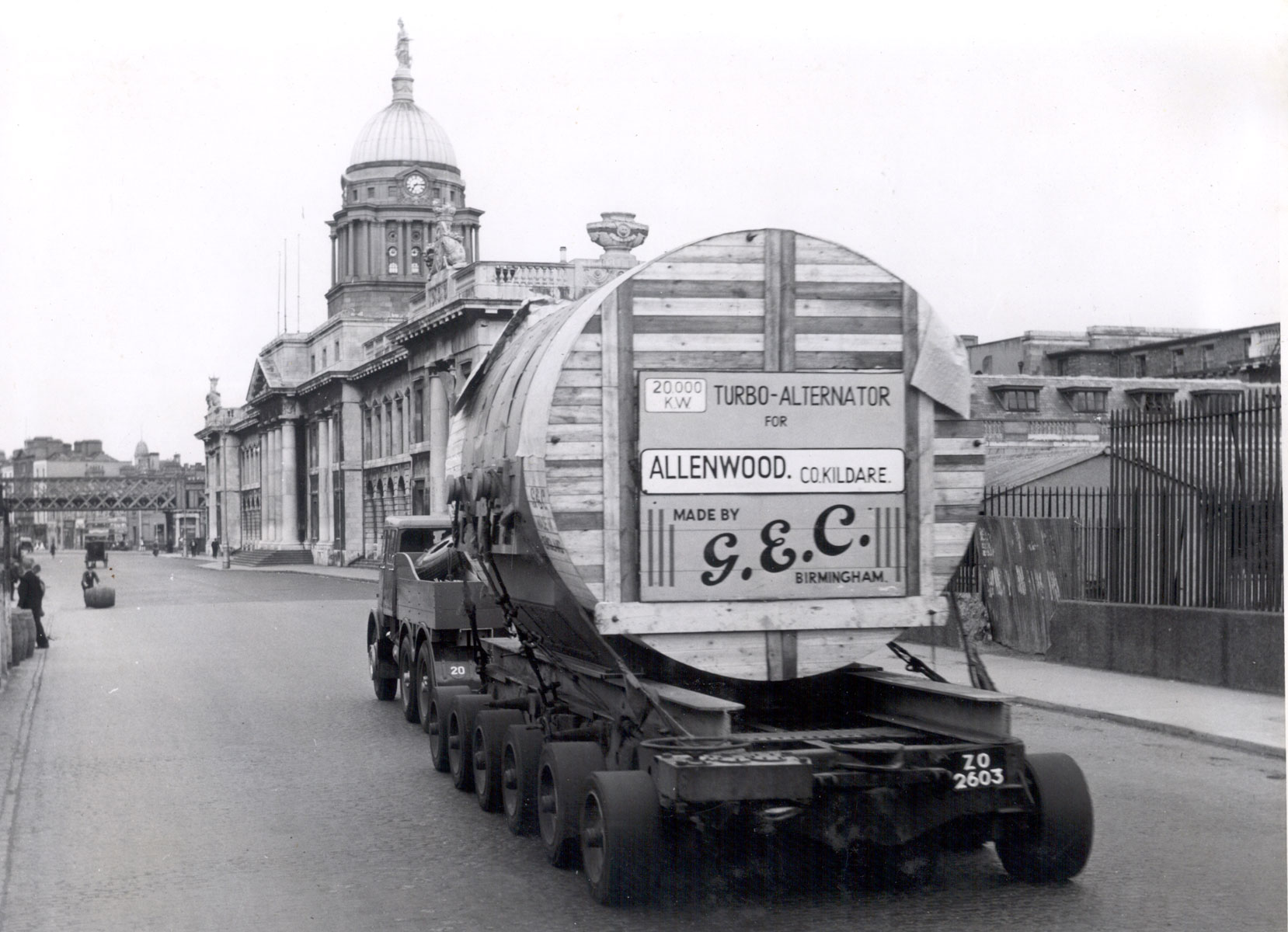 Black-and-white photograph showing a heavy transport vehicle carrying a large crated electrical generator component through a city street.