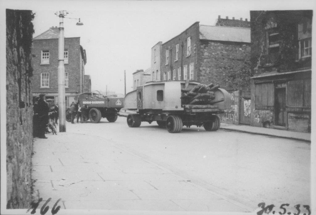 Black-and-white photograph showing a truck and transport trailer carrying large machinery through an urban street, with several people standing on the footpath and buildings on either side of the road.