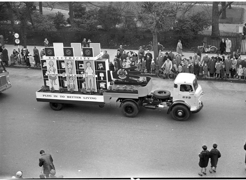 Black-and-white photograph showing an ESB promotional parade float mounted on a lorry, displaying messages about the benefits of electricity while spectators watch from the roadside.
